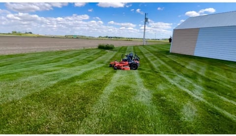 Red riding mower cutting diagonal lines through a large green lawn with farm buildings and fields visible in the background
