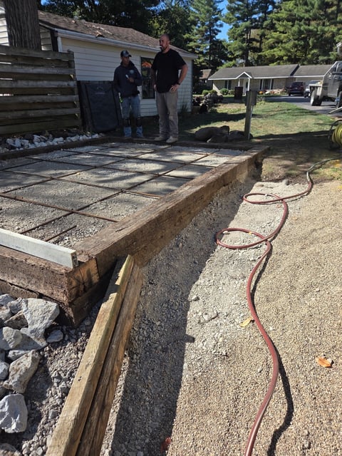 Construction site showing concrete foundation with wooden frame, two workers in background, and coiled red hose in foreground at residential property