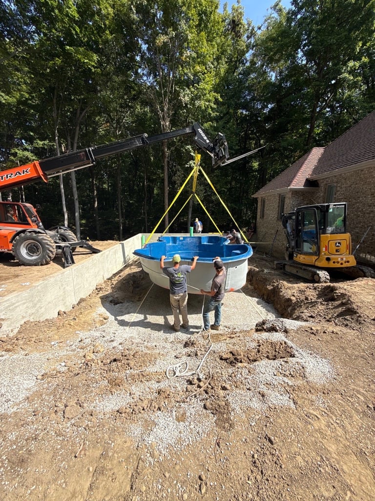 Two workers guide a blue pool being lowered by a telehandler on a residential construction site surrounded by heavy equipment