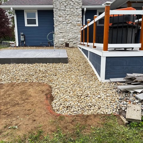 Backyard landscaping project showing a house with blue siding, stone chimney, wooden deck with metal railings, gravel and pavers for ground coverage