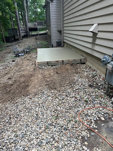Construction site beside gray house with exposed concrete foundation and gravel ground, wooden deck visible in background