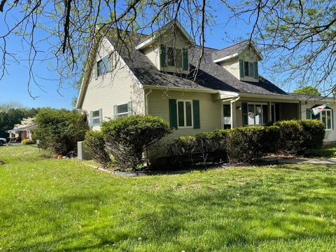 Cream-colored farmhouse with dormer windows, dark roof, green shutters, surrounded by manicured shrubs and lawn