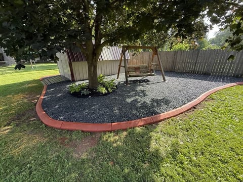 Backyard play area with wooden arbors and swings on gravel, bordered by red metal edging in green lawn