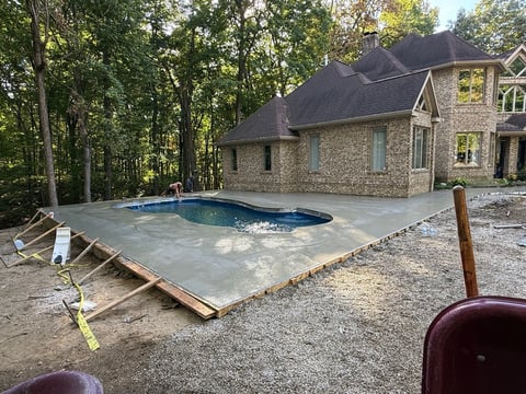 In-ground swimming pool under construction beside a two-story stone house with surrounding trees