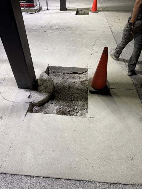 Concrete construction site with an open utility trench surrounded by red traffic cones and a black column pillar