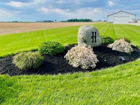 Large stone with cross on rural farmland surrounded by decorative plants and mulch, with barn in background