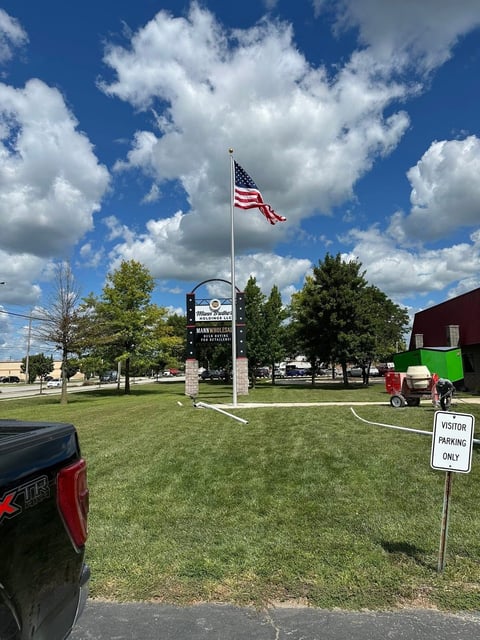 American flag on flagpole in grassy parking area with visitor parking sign and trees under blue sky with clouds