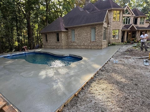 Luxury stone house with curved in-ground pool surrounded by concrete deck and wooded yard