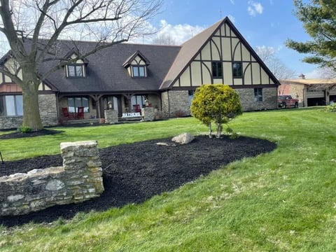 Tudor-style brick and stone home with manicured lawn, landscaped garden bed with young shrub, and bare trees in spring setting