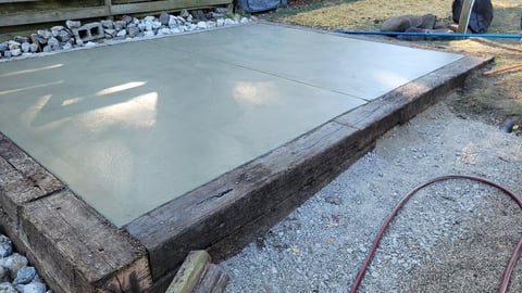Freshly poured concrete pad drying on gravel base with landscape border and garden hose nearby