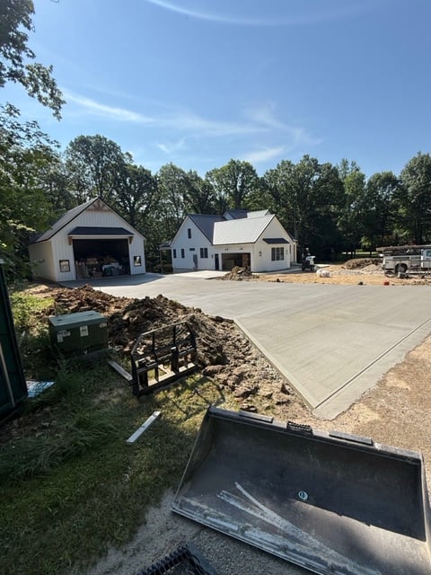 Construction site showing new residential home with garage, poured concrete driveway, and landscaping materials under clear blue sky