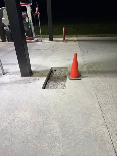Red traffic cone placed near an open square hole in concrete pavement at a service station at night