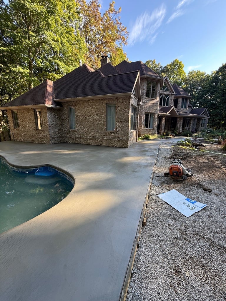 Large stone house with dark roof surrounded by autumn trees, featuring a new concrete patio and empty pool in foreground under construction