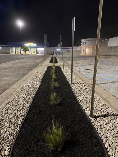 Night view of empty shopping center parking lot with landscaped median strip featuring small plants and metal posts
