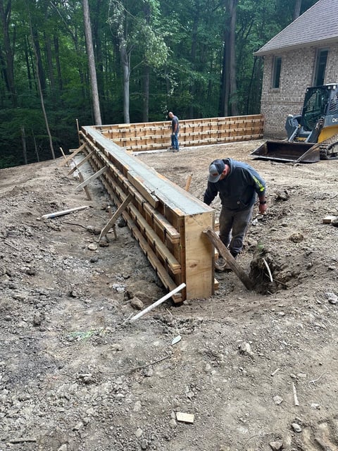 Construction worker in vest building wooden retaining wall on dirt site with brick house and heavy machinery in background