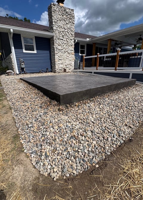 Modern home exterior with dark concrete patio, gravel landscaping, stone chimney, and covered deck area
