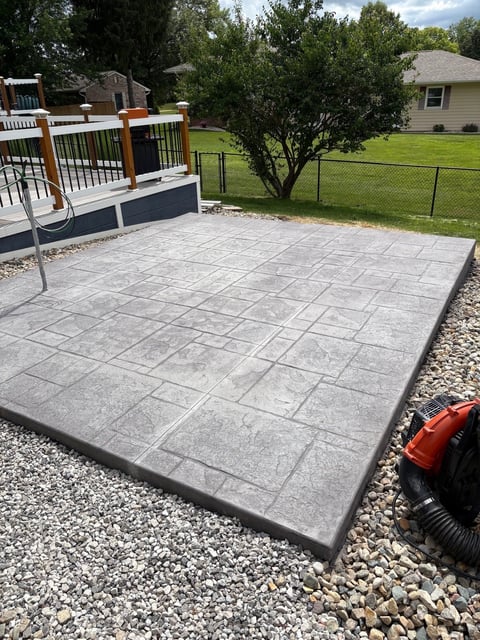 Gray patterned concrete patio installed in backyard with raised deck, gravel border, lawn and trees in background