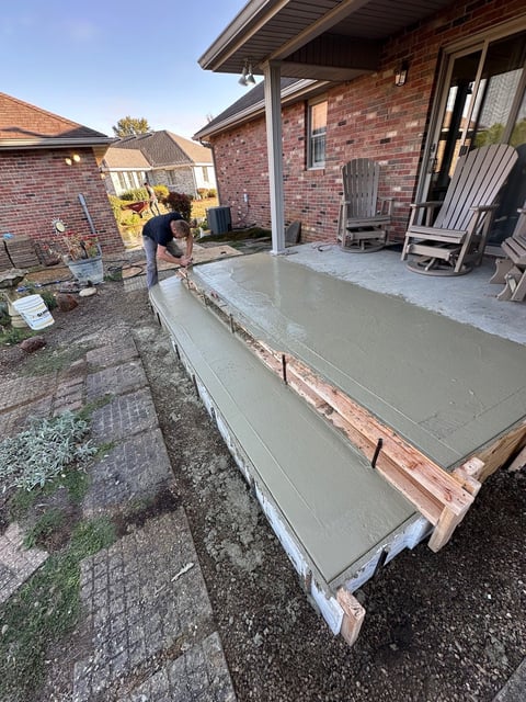 Worker smoothing wet concrete on a residential patio extension next to a brick house during construction