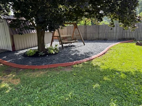 Backyard with green lawn, wooden swing set, large tree, gravel area with red border edging, and wooden fence