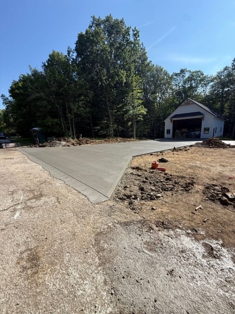 Newly poured concrete driveway next to a white garage with dark roof, surrounded by trees and dirt landscaping