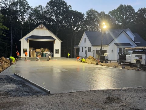 Freshly poured concrete driveway at night with white garage and house, construction workers present, illuminated by work lights